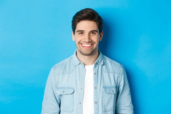 Nær-up av en ung suksessfull mann som smiler til kameraet mens han står i vanlige klær mot blå bakgrunn – stockfoto