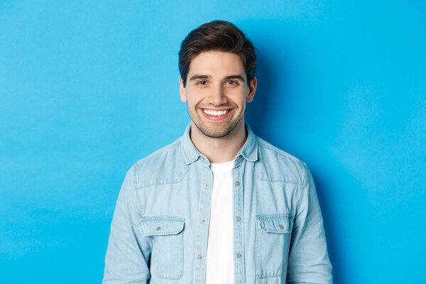 Close-up of young successful man smiling at camera, standing in casual outfit against blue background
