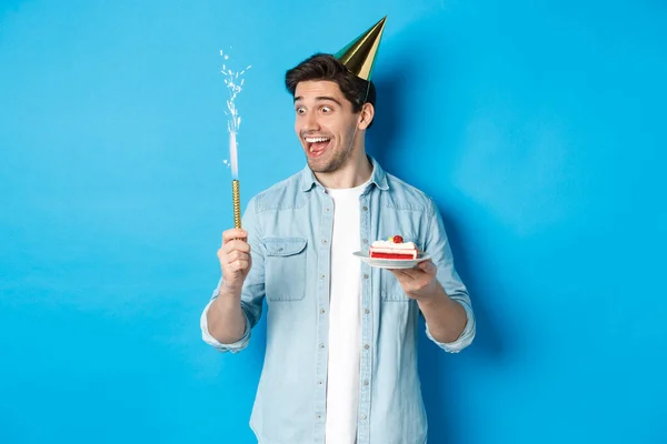 Gelukkige jongeman vieren verjaardag in feesthoed, houden b-day cake en glimlachen, staande over blauwe achtergrond — Stockfoto