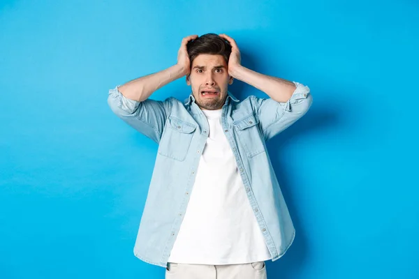 Изображение человека в панике, держащегося за голову, выглядящего расстроенным и встревоженным, стоящего на синем фоне — стоковое фото