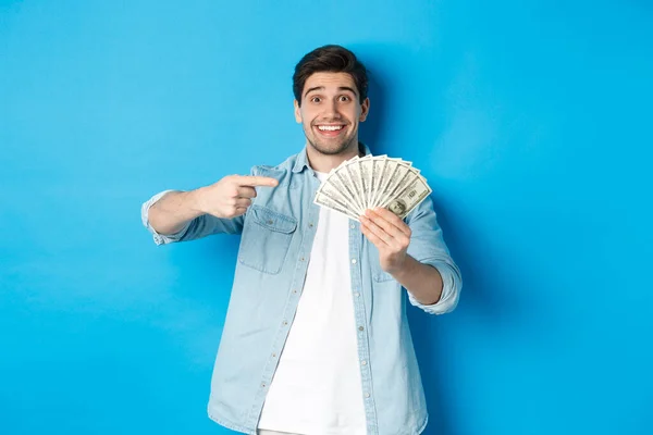Sorprendido hombre sonriente con ropa casual, señalando con los dedos al dinero, de pie sobre el fondo azul — Foto de Stock
