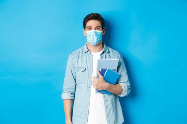 Educação, covid-19 e distanciamento social. Guy estudante em máscara médica olhando feliz, segurando cadernos, de pé sobre fundo azul — Fotografia de Stock