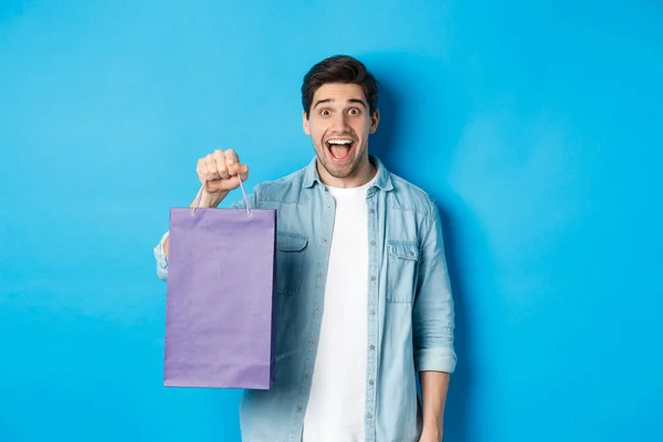 Concepto de compras, vacaciones y estilo de vida. Emocionado chico guapo sosteniendo bolsa de papel con regalo y mirando feliz, de pie sobre fondo azul — Foto de Stock