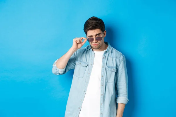 Homem macho confiante colocar em óculos de sol, olhando fresco e atrevida, de pé sobre fundo azul — Fotografia de Stock
