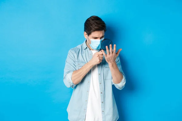 Conceito de coronavírus, distanciamento social e pandemia. Homem de máscara médica olhando para a palma da mão através de lupa, de pé sobre fundo azul — Fotografia de Stock