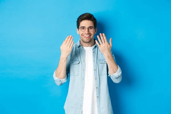 Joven guapo en gafas mostrando las manos de manicura y sonriendo, cuidando las uñas, de pie sobre fondo azul — Foto de Stock