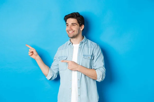 Knappe volwassen man introduceren product, kijken en wijzen vingers links, het bevorderen van iets tegen blauwe achtergrond — Stockfoto