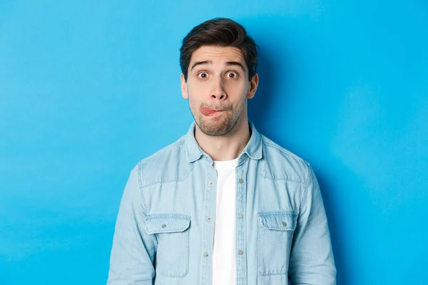 Close-up de jovem fazendo expressões engraçadas, mostrando a língua e olhando para a câmera, de pé sobre o fundo azul — Fotografia de Stock