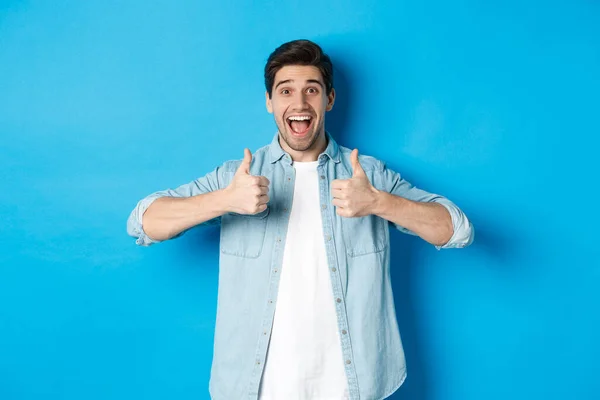 Hombre adulto sonriente mostrando los pulgares hacia arriba con la cara excitada, como algo impresionante, aprobando el producto, de pie sobre el fondo azul —  Fotos de Stock