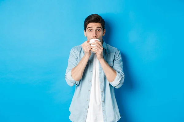 Мужчина выглядит взволнованным и потягивая чай или кофе из белой кружки, стоя на синем фоне — стоковое фото