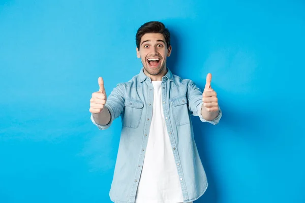 Glimlachende zelfverzekerde man die duimen laat zien met opgewonden gezicht, als iets geweldigs, het goedkeuren van product, staande tegen een blauwe achtergrond — Stockfoto