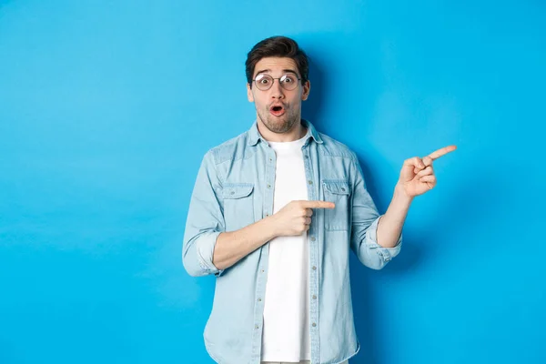 Hombre sorprendido en gafas apuntando a la derecha en el espacio de copia, mostrando oferta promocional sobre fondo azul, de pie sobre fondo azul — Foto de Stock