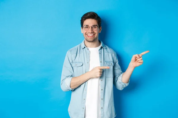 Knappe blanke man model in bril, wijzende vingers recht op uw logo, tonen kopieerruimte, staande over blauwe achtergrond — Stockfoto