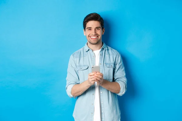 Beau barbu en tenue décontractée souriant à la caméra, vérifiant smartphone, debout sur fond bleu — Photo