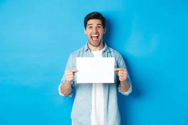 Hombre atractivo feliz mostrando pedazo de papel para su signo de logotipo, de pie sorprendido contra el fondo azul — Foto de Stock
