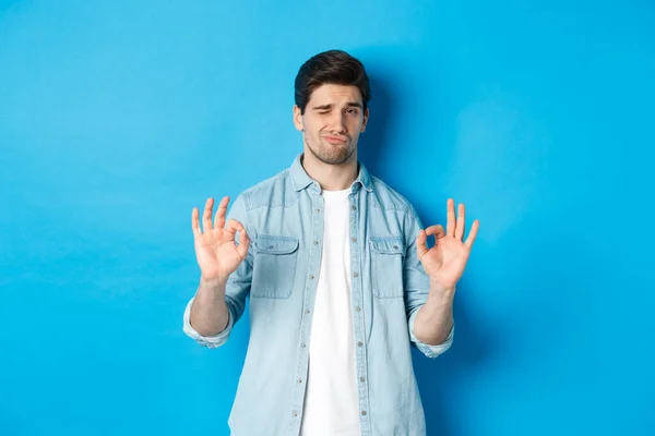 Tevreden knappe man met een-ok tekenen en kijken blij, goedkeuring van iets goeds, staande tegen een blauwe achtergrond — Stockfoto