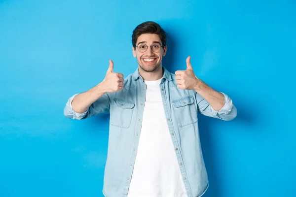 Atractivo joven con gafas y ropa casual, mostrando los pulgares hacia arriba en aprobación, como algo, de pie sobre fondo azul — Foto de Stock