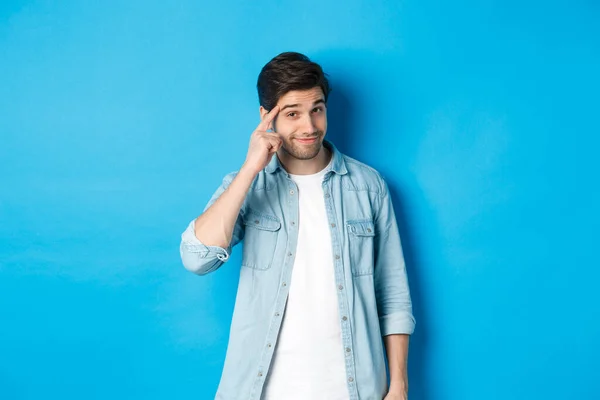 Jonge attente man wijst naar het hoofd, vraagt om erover na te denken, geeft een hint, staat in casual kleding over blauwe achtergrond — Stockfoto