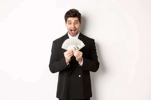 Imagen del guapo barbudo en traje negro, mirando el dinero con emoción, de pie sobre fondo blanco — Foto de Stock