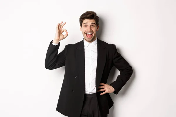 Concepto de fiesta de año nuevo, celebración y estilo de vida. Retrato de chico guapo complacido en traje negro, elogiar algo bueno, mostrando signo bien en la aprobación, de pie sobre fondo blanco —  Fotos de Stock