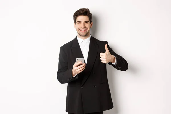 Handsome guy in trendy suit making compliment, showing thumbs-up while using mobile phone, recommending an app or online shop, standing over white background — Stock Photo, Image