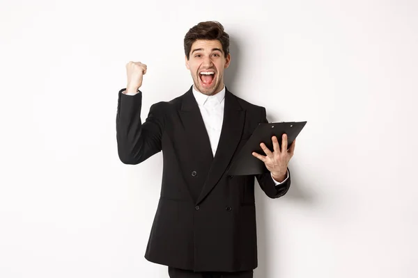 Portrait of excited handsome businessman in black suit, holding clipboard and making fist pump, achieve goal and rejoicing, standing over white background — Stock Photo, Image