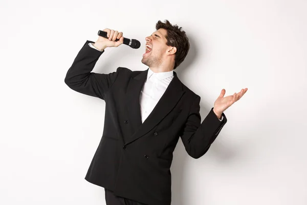 Retrato de homem bonito cantando uma canção com paixão, de pé em terno preto, segurando microfone e performning, posando sobre fundo branco — Fotografia de Stock