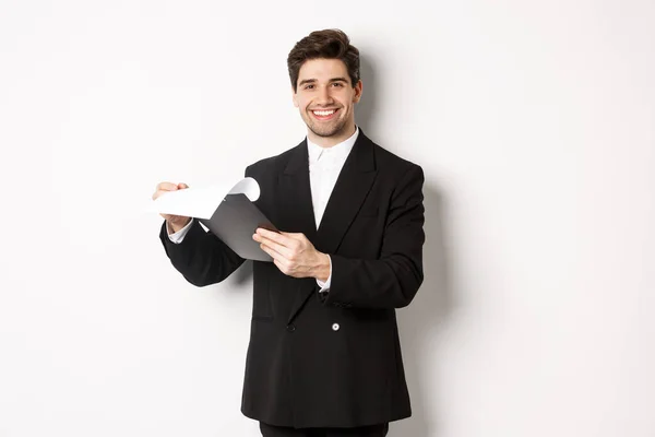 Imagen de empresario confiado en traje mirando documentos, sujetando el portapapeles y sonriendo complacido, de pie sobre fondo blanco — Foto de Stock