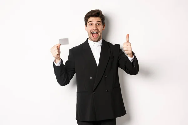 Bild av snygg affärsman i svart kostym, visar tummen upp och kreditkort, rekommendera bank, står mot vit bakgrund — Stockfoto
