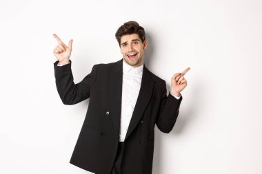 Yakışıklı adam dans ediyor ve siyah takım elbise içinde parti yapıyor, parmaklarını yana doğru gösteriyor, iki reklam pankartı gösteriyor, beyaz arka planda duruyor.