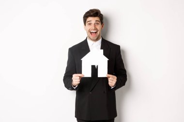 Siyah takım elbiseli yakışıklı bir emlakçının ev yapımı maketini göstermesi, şaşkın görünmesi, ev satması, beyaz arka planda durması.