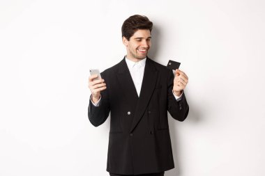 Çevrimiçi alışveriş yapan kendine güvenen yakışıklı iş adamının portresi, elinde kredi kartı ve akıllı telefon, beyaz arka planda duruyor.