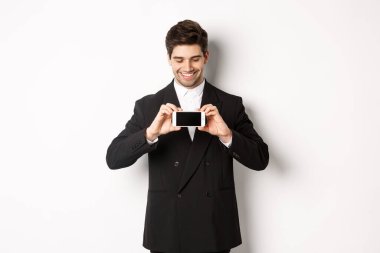 Siyah takım elbiseli yakışıklı sakallı adamın portresi, size akıllı telefon ekranını gösteriyor ve gülümsüyor, beyaz arka planda duruyor.