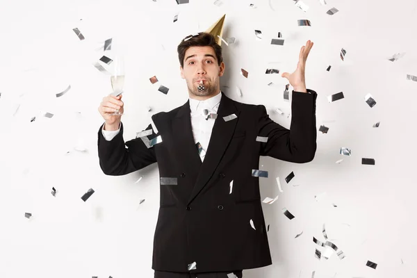 Handsome party guy in black suit having fun, celebrating new year, blowing whistle and drinking champagne while confetti falling, wishing happy holidays, standing against white background