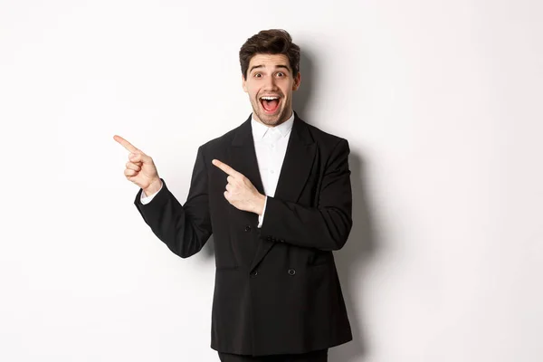Portrait of excited and happy man in suit showing advertisement, pointing fingers at upper left corner and smiling amazed, standing over white background — Stock Photo, Image