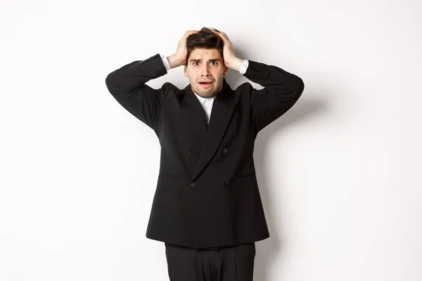 Hombre de negocios frustrado y preocupado en traje negro, asustado como mirando a los problemas, tomados de la mano en la cabeza alarmado, de pie sobre fondo blanco — Foto de Stock