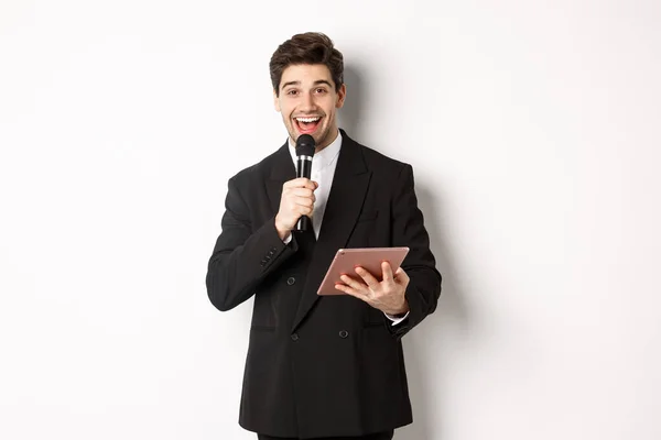 Imagen de un guapo artista masculino, dando un discurso, entretener a la gente en la fiesta, sosteniendo micrófono y tableta digital, de pie sobre fondo blanco — Foto de Stock