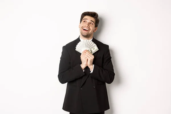Bilde av en kjekk, drømmende mann i svart dress, som holder penger og ser opp mot venstre hjørne, tenker på shopping, står over hvit bakgrunn – stockfoto