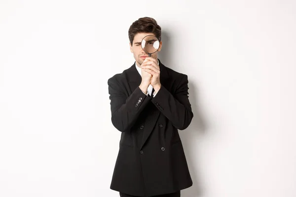 Imagen de hombre de negocios serio en traje negro de moda, mirando a través de lupa, buscando empleados, de pie sobre fondo blanco — Foto de Stock