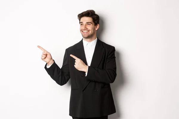 Porträtt av framgångsrik stilig man i kostym, pekar och tittar vänster med glada leende, visar promo banner, står över vit bakgrund — Stockfoto