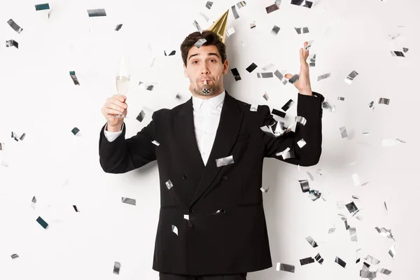 Handsome party guy in black suit having fun, celebrating new year, blowing whistle and drinking champagne while confetti falling, wishing happy holidays, standing against white background