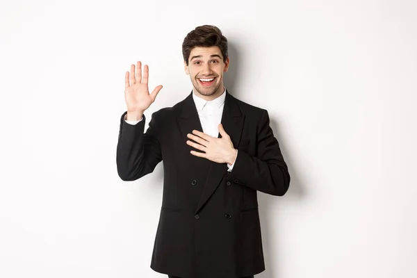 Wizerunek przystojnego uśmiechniętego faceta mówiącego prawdę, podnoszącego rękę, by coś obiecać, stojącego nad białym tłem w czarnym garniturze — Zdjęcie stockowe