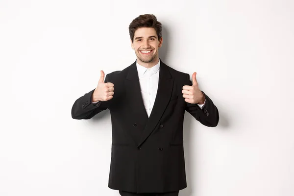 Concept of new year party, celebration and lifestyle. Image of attractive happy businessman in formal suit, showing thumbs-up and smiling, like and approve, standing over white background — Stock Photo, Image