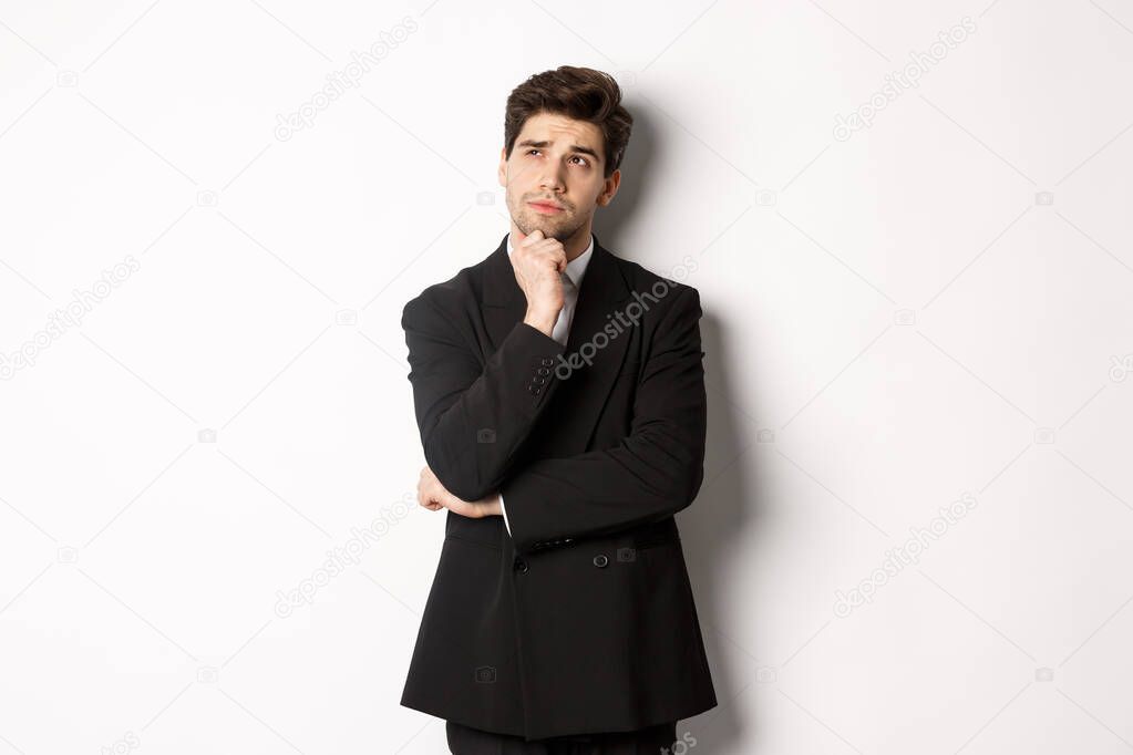 Portrait of handsome and stylish man in suit, thinking and looking at upper left corner, imaging something about christmas holidays, standing over white background.
