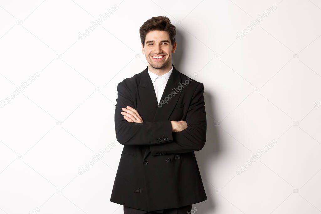 Handsome and confident bearded man, standing in formal suit with hands crossed on chest, smiling satisfied, attend party or work event, standing over white background.