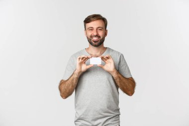 Gri tişörtlü neşeli beyaz adam, kredi kartı gösteriyor ve gülümsüyor, alışveriş için heyecanlı, beyaz arka planda duruyor.