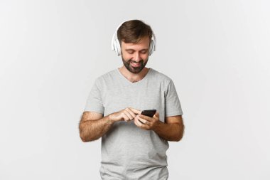 Çekici modern adamın portresi gülümseyen, cep telefonuna bakan, kulaklıkla müzik dinleyen, beyaz arka planda duran