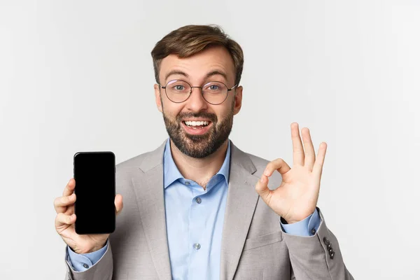 Close-up of excited businessman in gray suit and glasses, recommending good app, showing mobile phone screen and okay sign, standing over white background — Stock Photo, Image