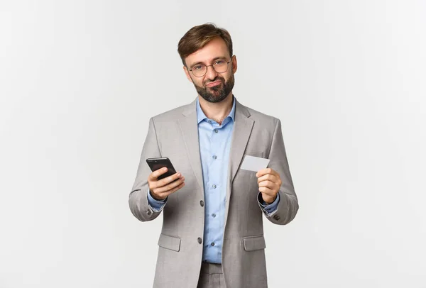 Portrait of skeptical businessman in glasses and grey suit, grimacing disappointed, holding mobile phone and credit card, standing over white background — Stock Photo, Image