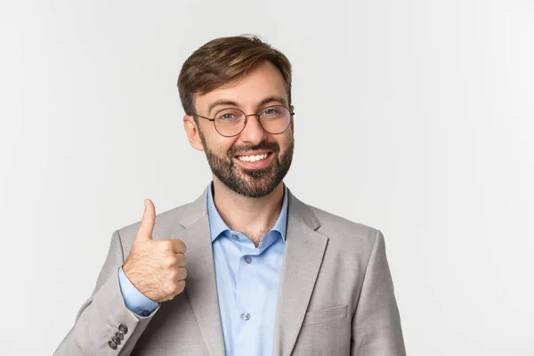 Close-up de homem barbudo bonito em óculos e terno cinza, mostrando aprovação polegar-up, como algo, recomendar produto ou marca, de pé sobre fundo branco — Fotografia de Stock